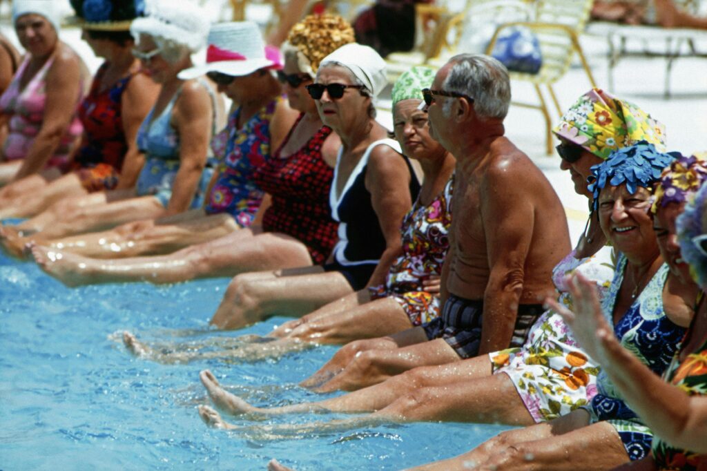 “Men and women in their sixties sitting poolside in swimwear, smiling and enjoying time together as a group.