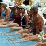 “Men and women in their sixties sitting poolside in swimwear, smiling and enjoying time together as a group.